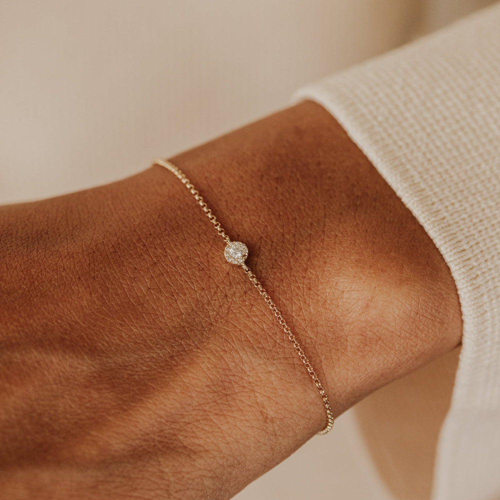 Gold bracelet with a small round pendant on a wrist against a neutral background.