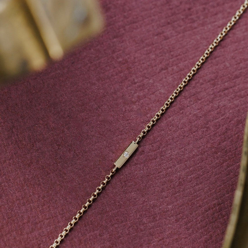 A close-up of a delicate gold chain bracelet with a round diamond set in a gold bar, laid diagonally on a textured dark purple fabric surface.