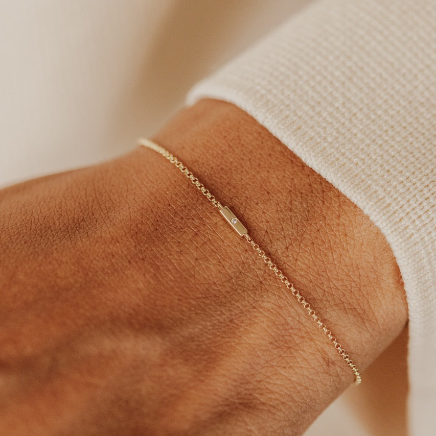 A delicate gold chain bracelet with a round natural diamond set in a gold bar, worn on a person's wrist over a white ribbed knit sleeve.