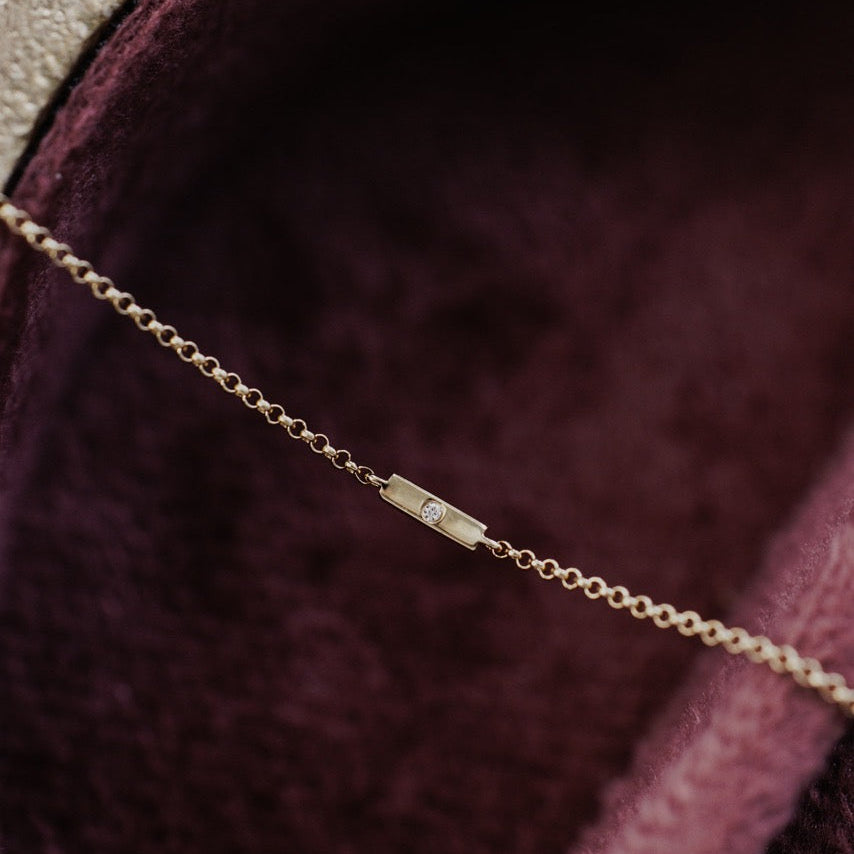 A close-up of a delicate gold chain bracelet with a round natural diamond set in a gold bar resting inside a plush burgundy velvet jewelry box.