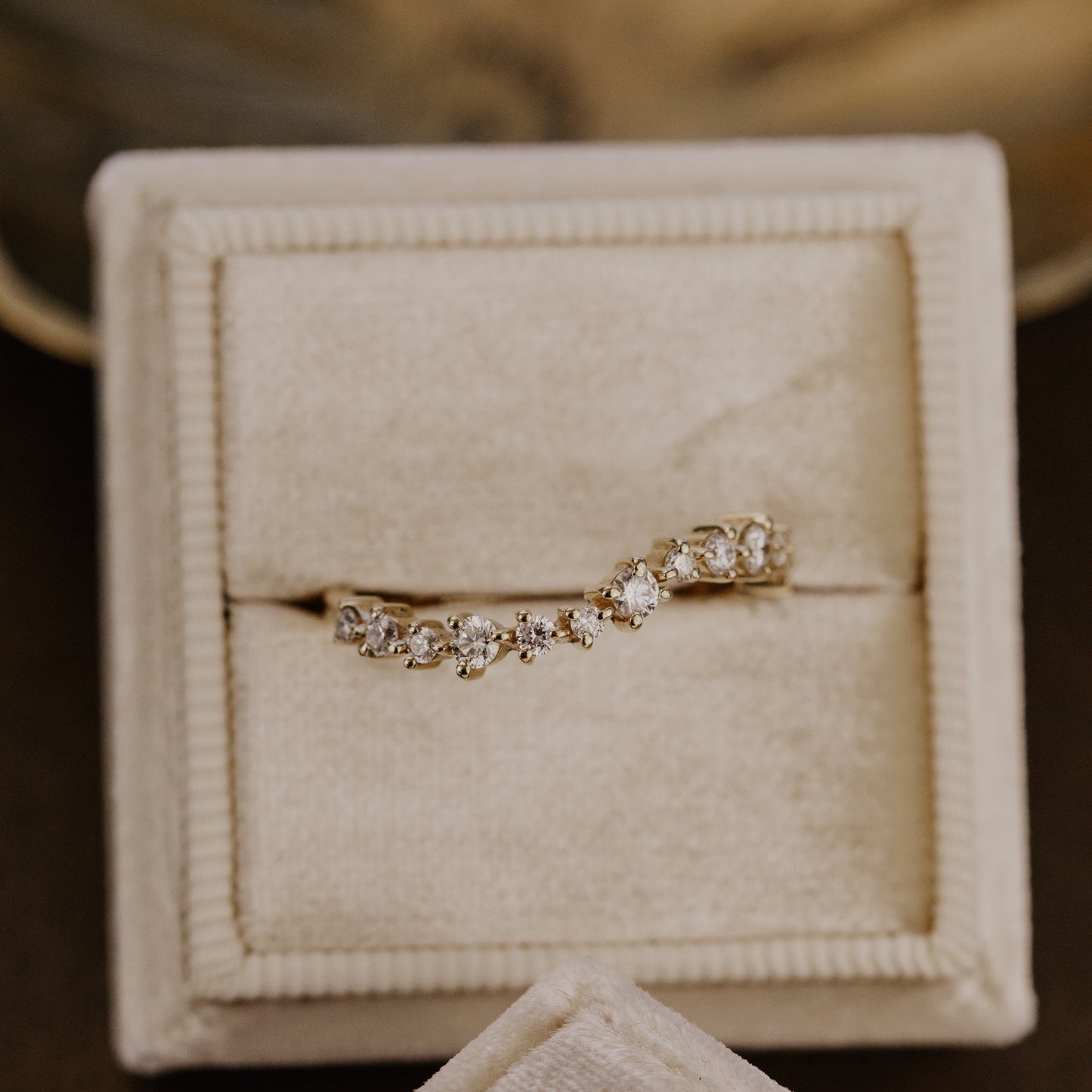 Gold wave-shaped wedding band with diamonds in a beige jewelry box.