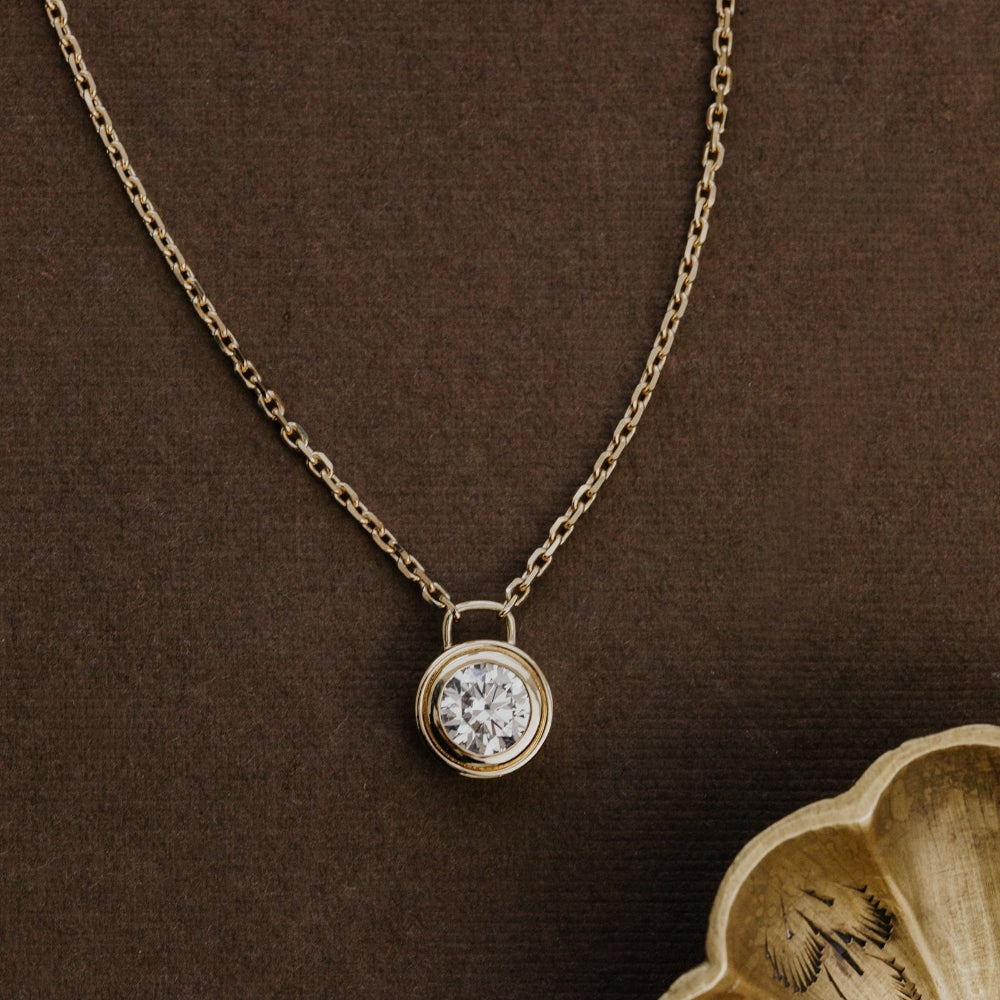 Gold necklace with a round lab grown diamond pendant on a brown background.