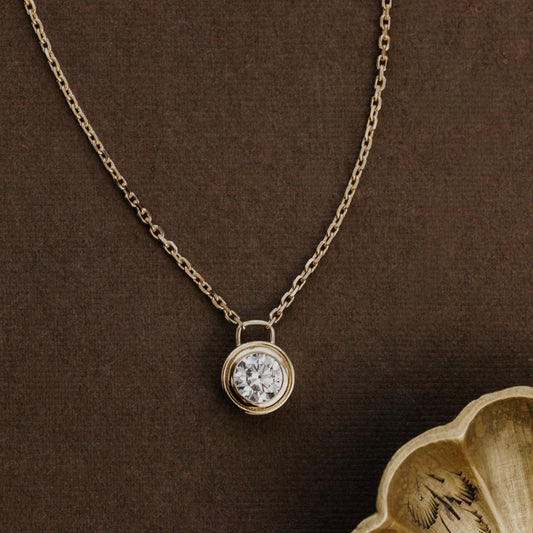Gold necklace with a round lab grown diamond pendant on a brown background.