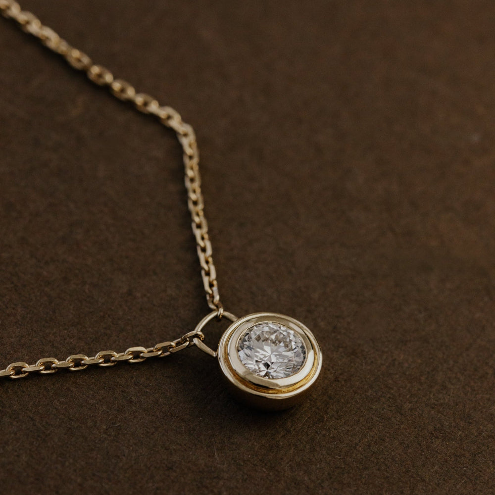 Gold necklace with a round lab grown diamond pendant on a brown textured background.