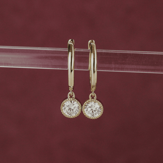 14k gold diamond earrings with a bezel design with huggie and dangle designs showcased on a clear bar in the center of the frame. 