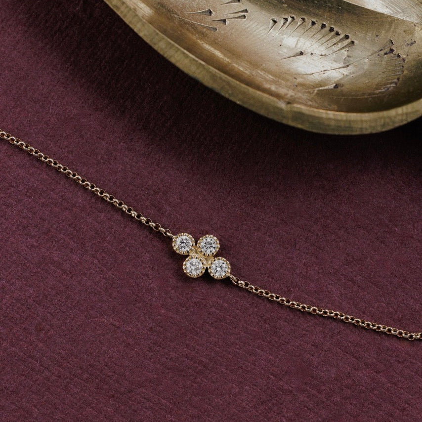 A delicate gold chain necklace featuring a diamond cluster displayed on a textured dark burgundy surface with a decorative gold tray in the background.