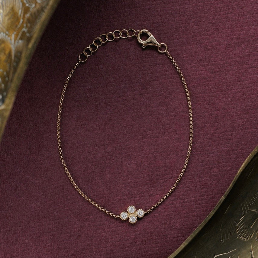 A delicate gold chain bracelet with a diamond cluster displayed on a textured dark purple fabric background with decorative metallic elements in the corners.
