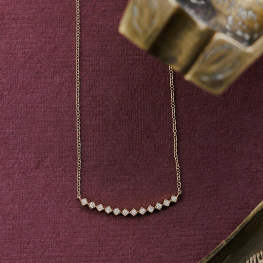 A delicate gold chain necklace with a curved row of small diamonds, displayed on a textured dark purple fabric background with decorative brass props.