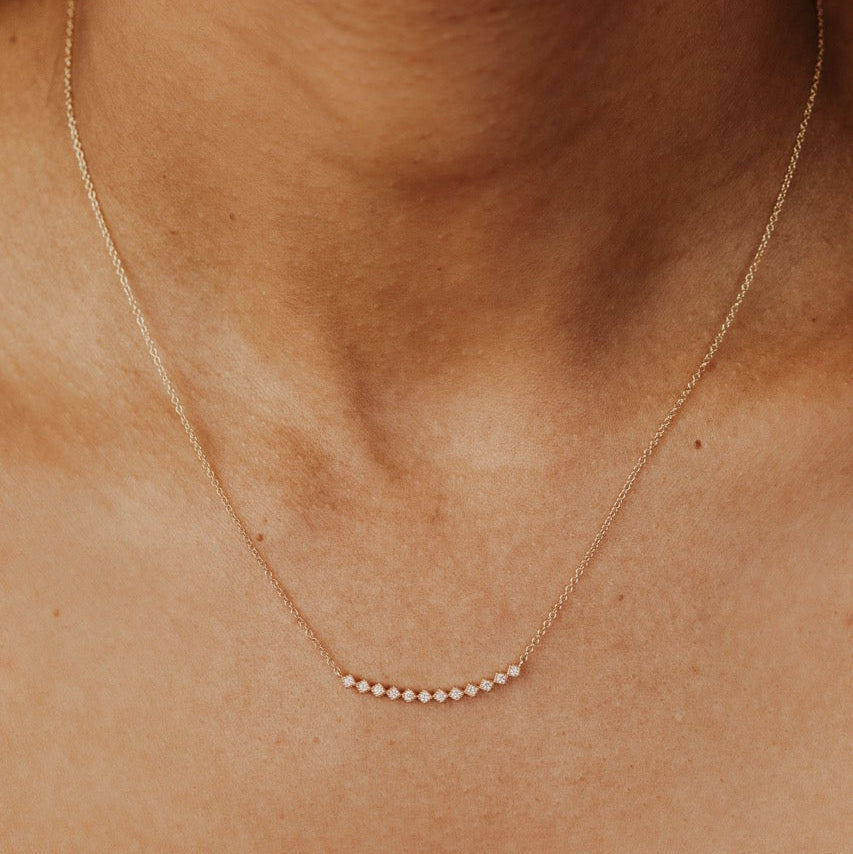 A delicate gold chain necklace with a curved row of small diamonds at the center, worn on a person's neck and upper chest against a dark background.