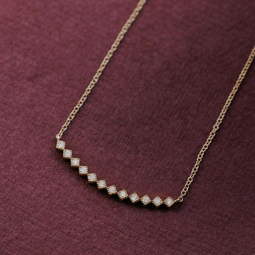 A delicate gold chain necklace featuring a curved row of small, sparkling diamonds, displayed on a textured dark purple fabric background.