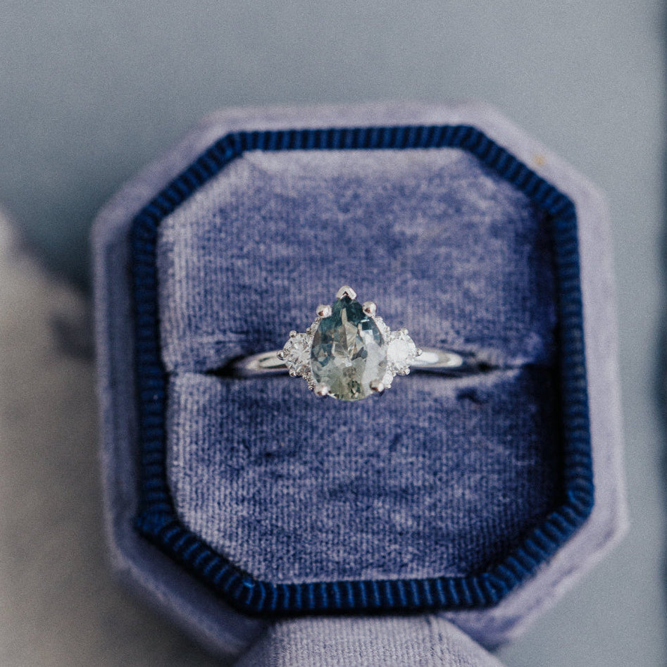 A pear-shaped light green sapphire ring with prong setting, accompanied by natural diamond side stones, displayed in a blue velvet ring box.