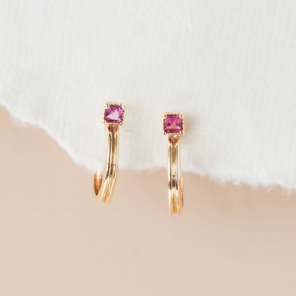 A pair of pink tourmaline earrings with thin textured gold hoops, displayed against a neutral background.