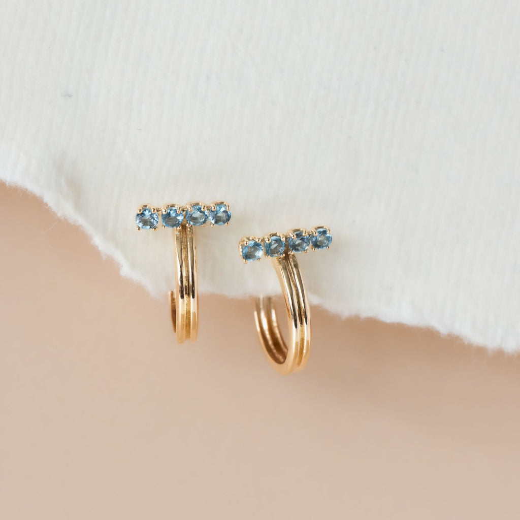 A pair of 14k yellow gold hoop earrings with blue topaz stones set on a bar, displayed against a white fabric backdrop.