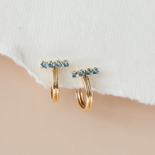 A pair of 14k yellow gold hoop earrings with blue topaz stones set on a bar, displayed against a white fabric backdrop.