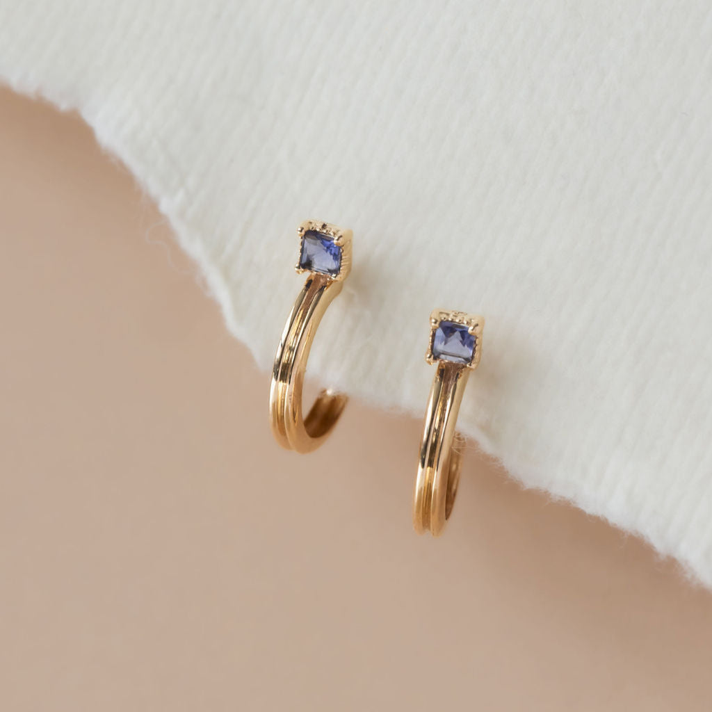 A pair of 14k yellow gold hoop earrings with a princess cut iolite stone set in prongs with a milgrain halo, displayed against a neutral background.