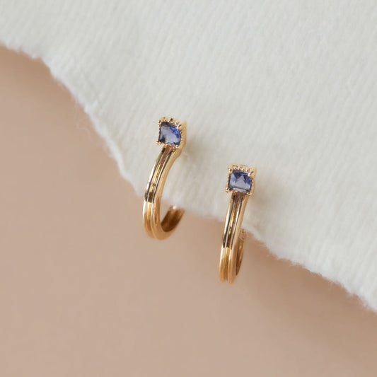 A pair of 14k yellow gold hoop earrings with a princess cut iolite stone set in prongs with a milgrain halo, displayed against a neutral background.