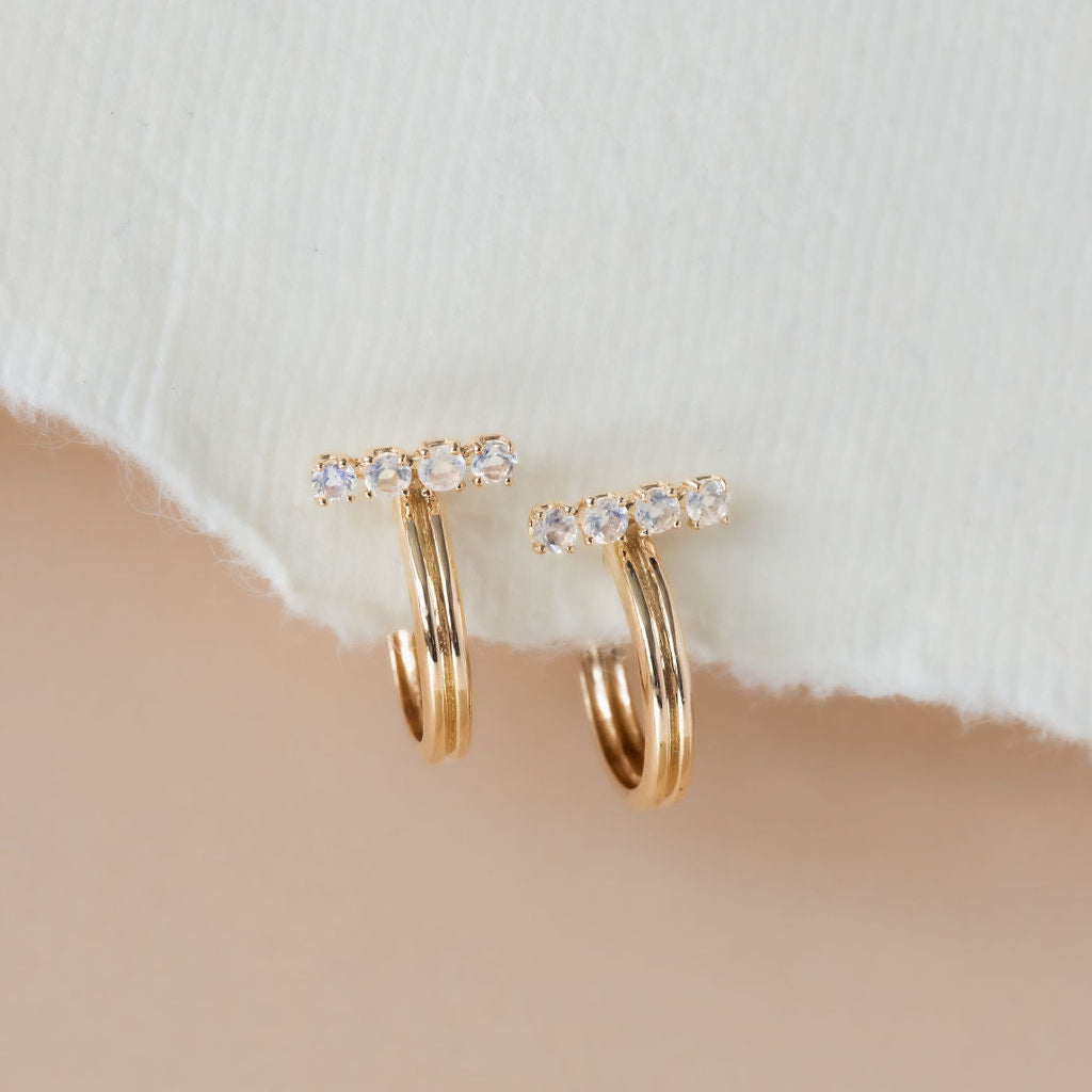 A pair of 14k yellow gold hoop earrings with round moonstone set on a bar, displayed against a neutral background.