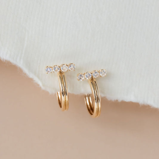 A pair of 14k yellow gold hoop earrings with round moonstone set on a bar, displayed against a neutral background.