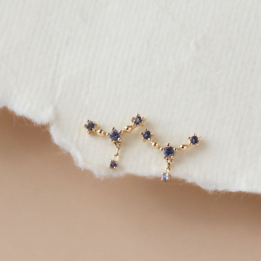 A pair of 14k yellow gold earrings featuring a curved bar design with iolite stones, displayed against a white fabric background.
