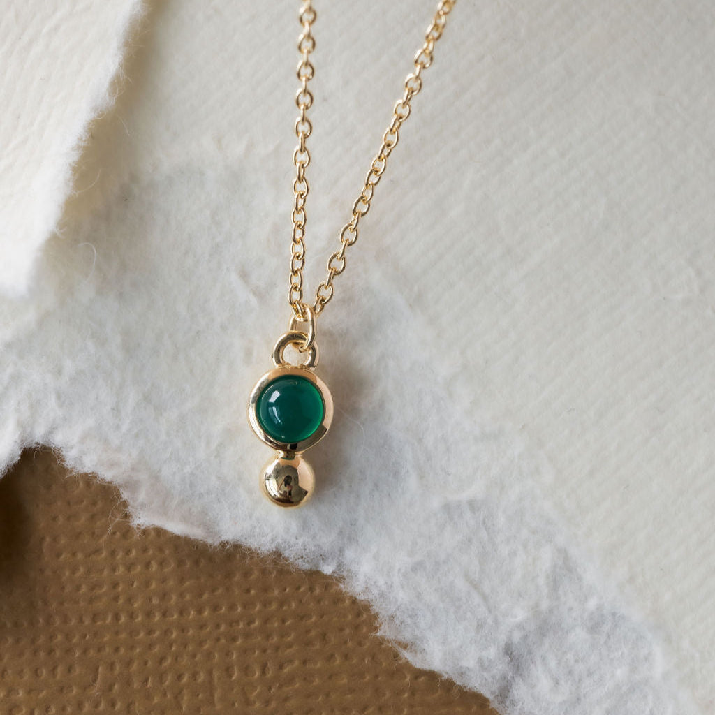 A gold necklace with a round green agate set in a bezel and a metal ball accent, displayed against a white and brown background.