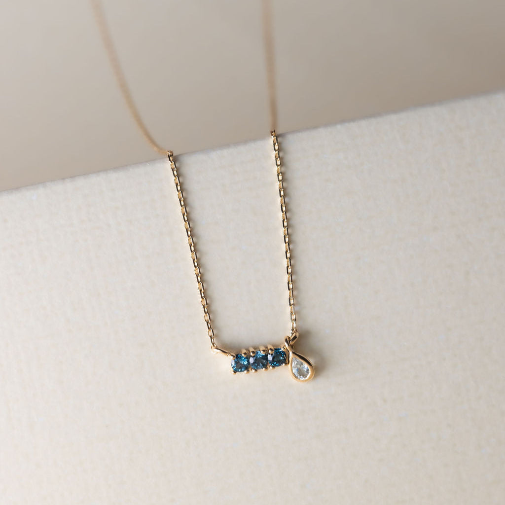 A 14k yellow gold necklace with a bar pendant set with blue topaz stones and a pear-shaped blue topaz dangle on an adjustable chain.
