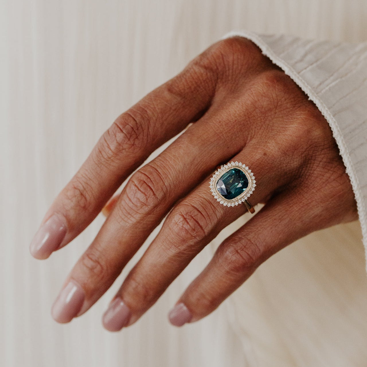 Hand wearing a ring with a diamond halo and a elongated cushion cut green Madagascar sapphire center stone on a neutral background