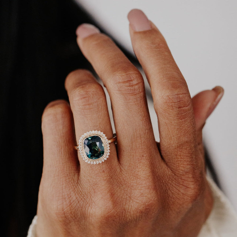 Hand wearing a diamond halo ring with a elongated cushion cut green Madagascar sapphire center stone  on a neutral background