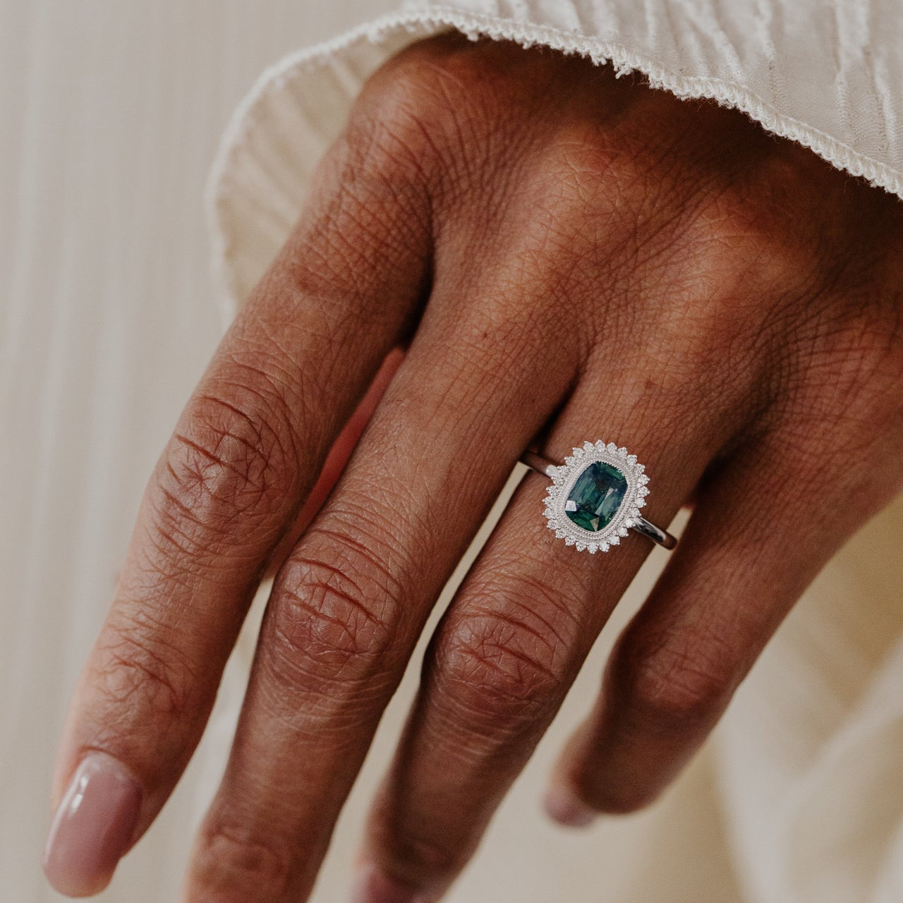 Hand wearing a engagement ring with a teal sapphire center stone and a diamond halo on a neutral background
