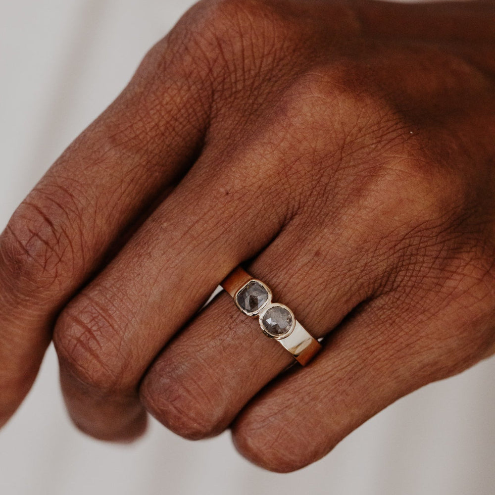 Hand wearing a yellow gold ring with two rustic diamond stones on a neutral background.