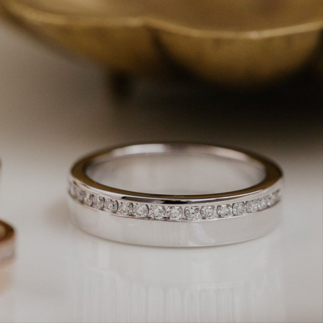 White gold ring with diamonds on a white surface with a blurred gold bowl in the background