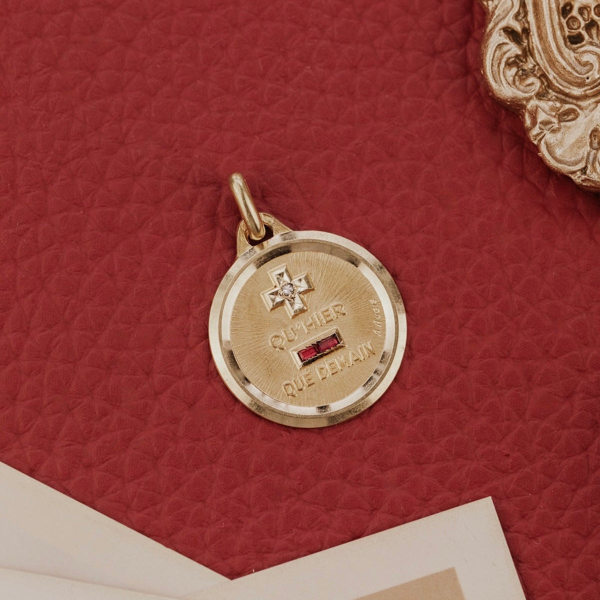 The Large 18k Yellow Gold Vintage French Love Token with Ruby Baguettes by A. Augis, featuring a round 18k yellow gold design with a knife-edge border, ruby baguettes, and the engraved phrase "qu'hier que demain," displayed on a deep red surface with an ornate gold frame.
