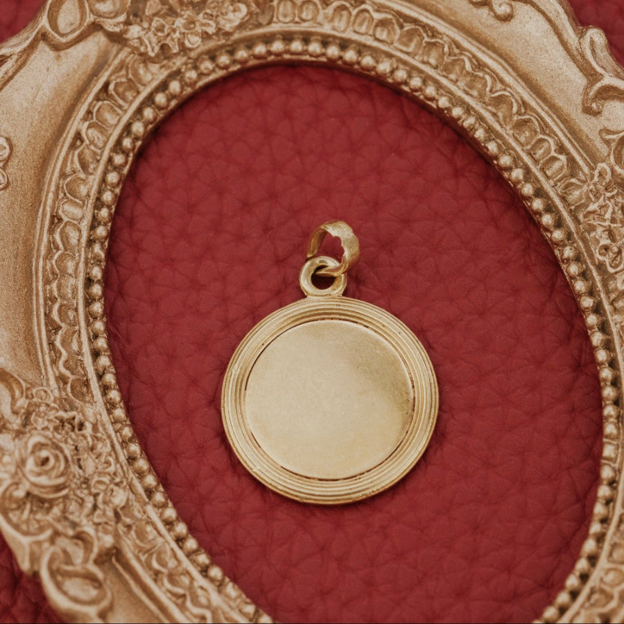 The Large 18k Yellow Gold Vintage Love Token with Diamonds by A. Augis, featuring a round design with a textured border, showcasing the back of the pendant displayed on a rich red surface.