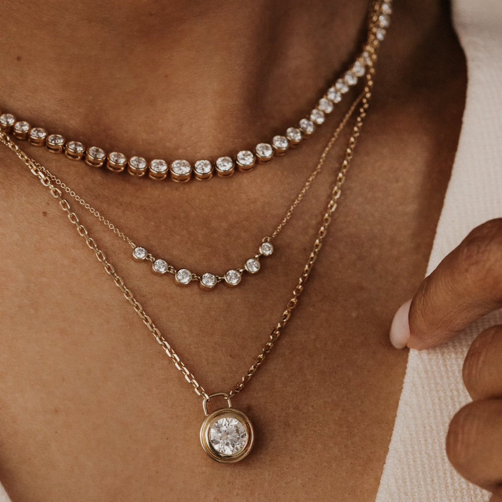 Close-up of a person wearing multiple diamond necklaces and rings on a neutral background,