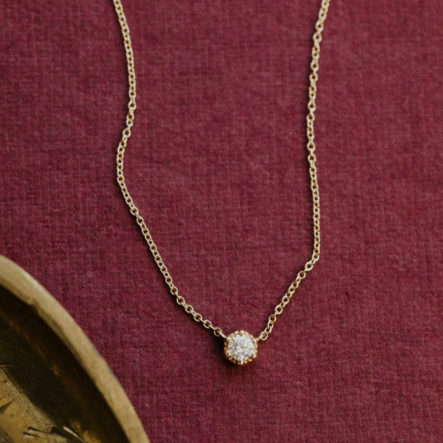 Gold necklace with a small round diamond pendant on a maroon fabric background