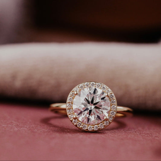 Gold ring with a large round diamond and smaller diamonds around on a pink surface.