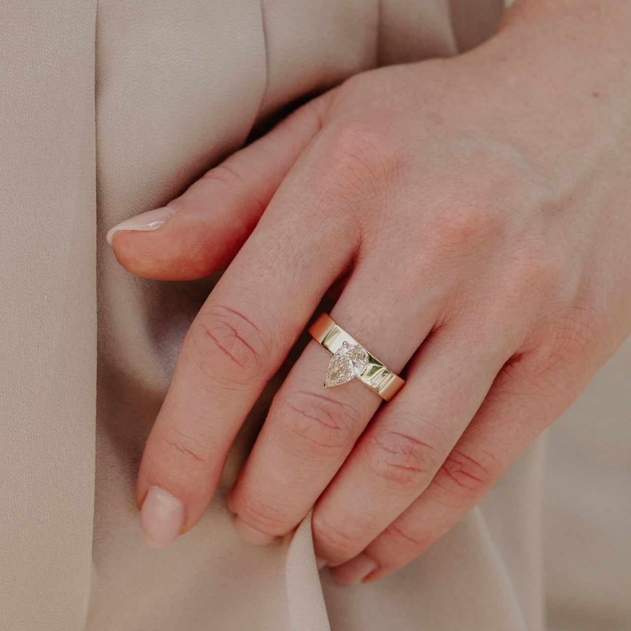 Hand wearing a gold ring with a pear shaped champagne diamond on a beige background