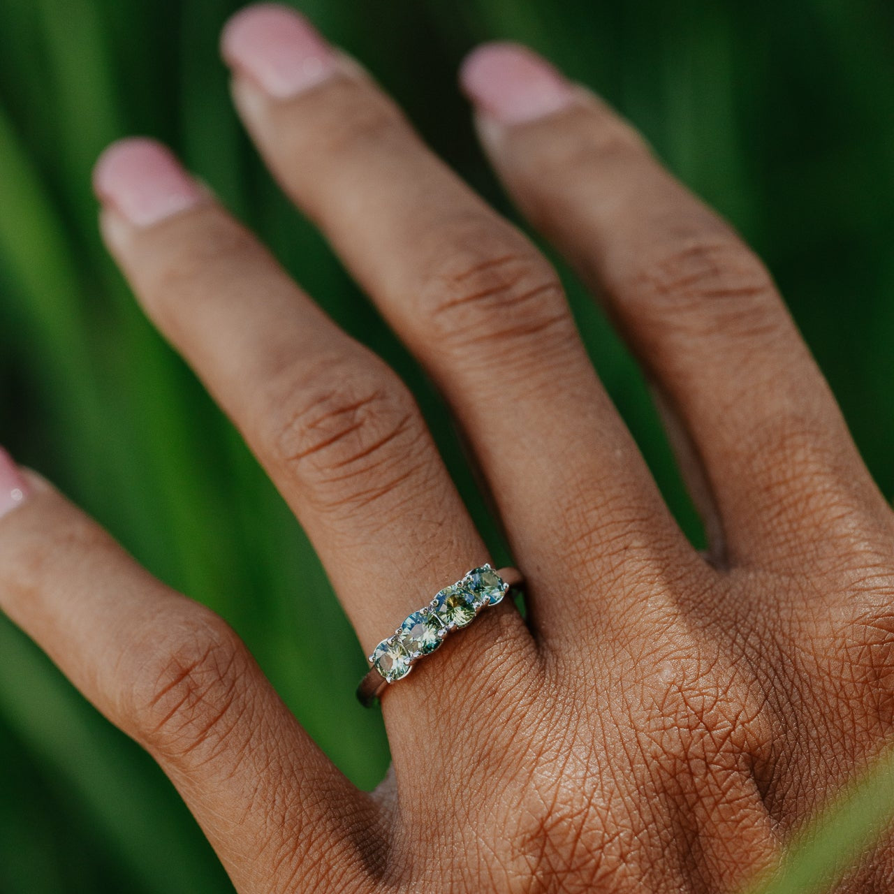 Hand wearing a ring with green and yellow sapphires against a blurred green background