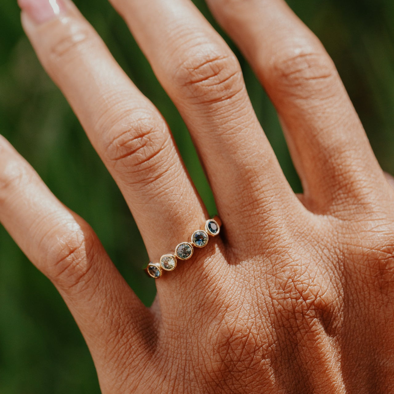 Hand wearing a gold ring with blue and green sapphires greenery in the background