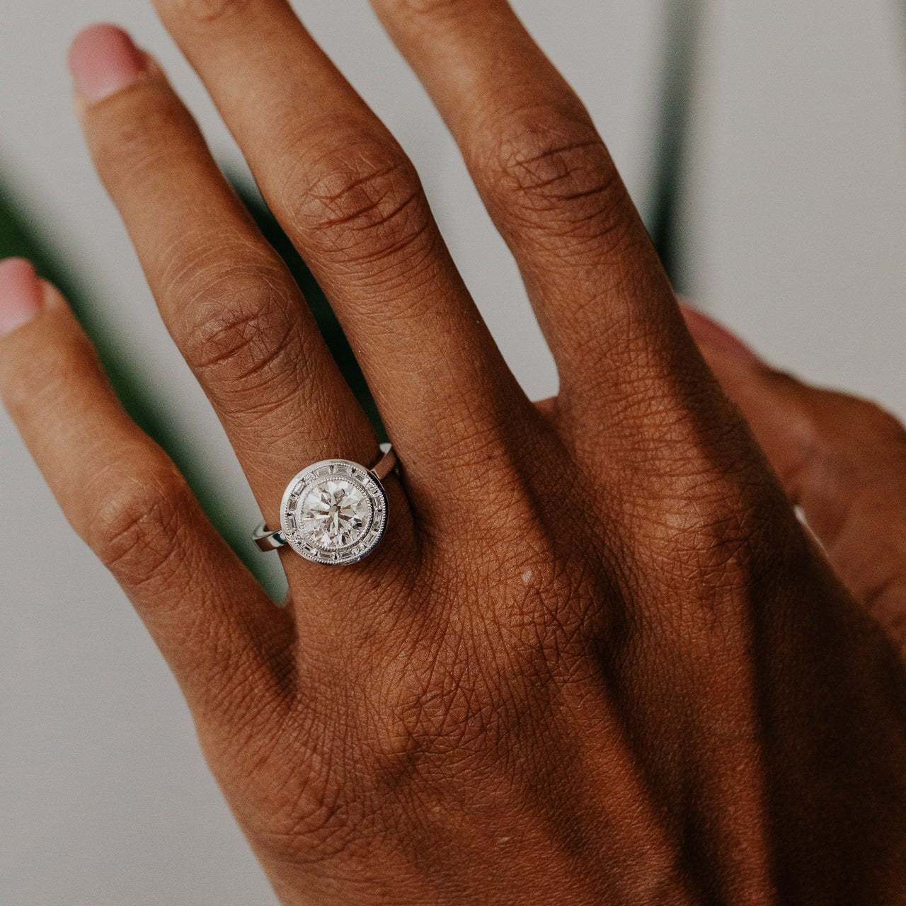 Hand wearing a white gold diamond halo ring with a round center diamond on a neutral background