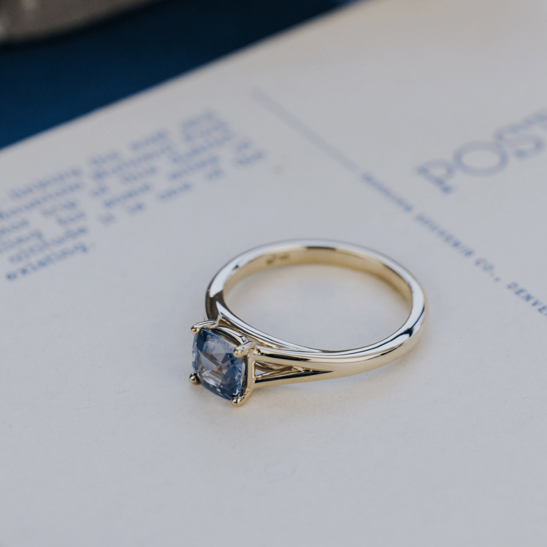A 14k gold ring featuring a side view of a blue Montana sapphire gemstone, displayed on a white surface with a soft background. 