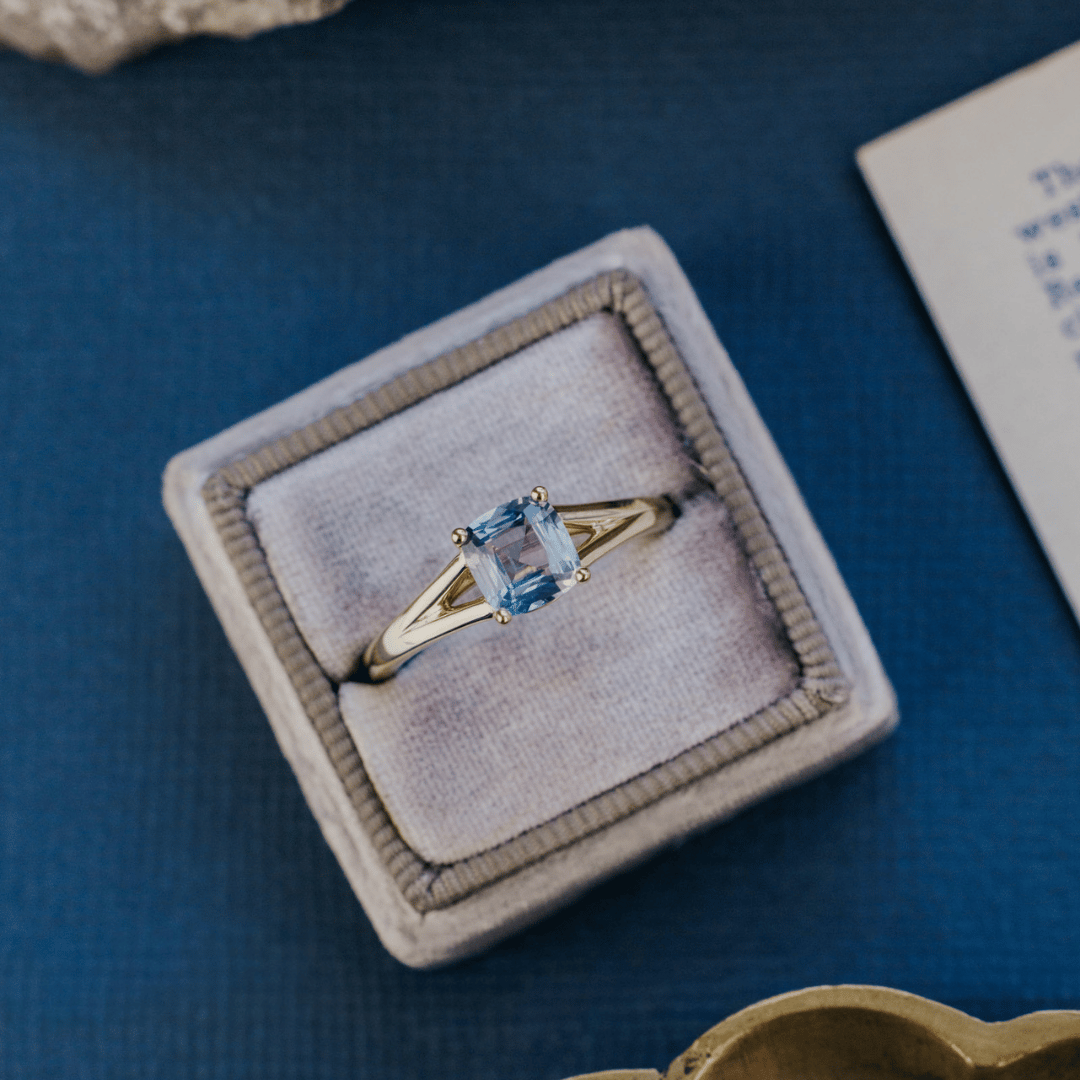 A 14k gold ring featuring a blue Montana sapphire gemstone, displayed in a ring box with a soft background. 