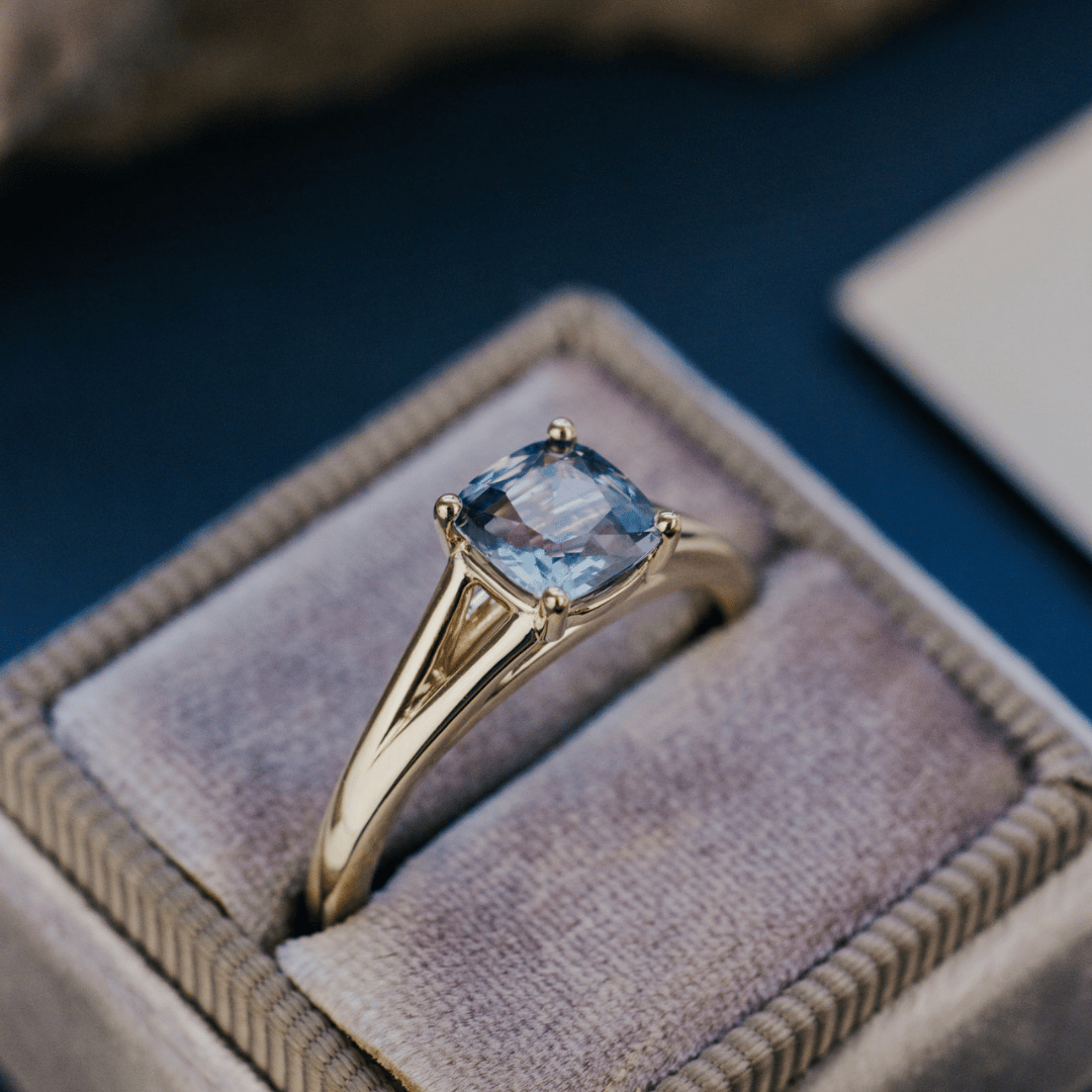 A 14k gold ring featuring a blue Montana sapphire gemstone, displayed in a ring box with a soft background. 