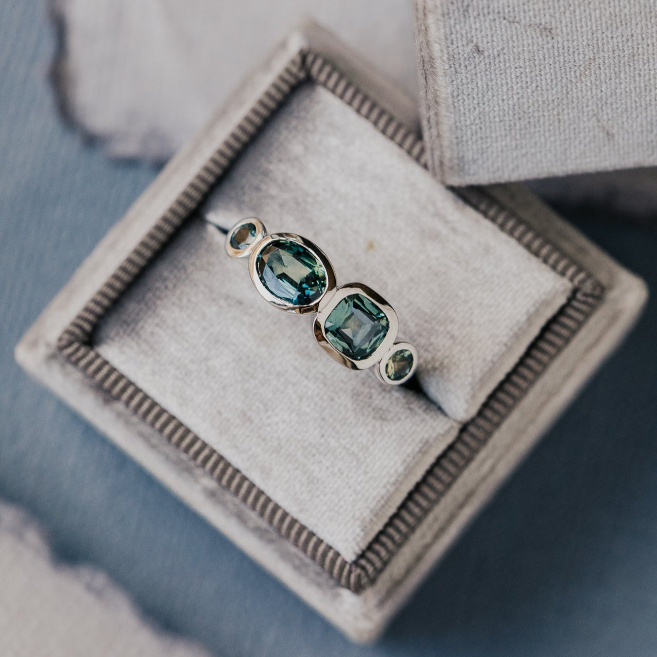White gold ring with green and blue sapphiresin a decorative box on a textured surface