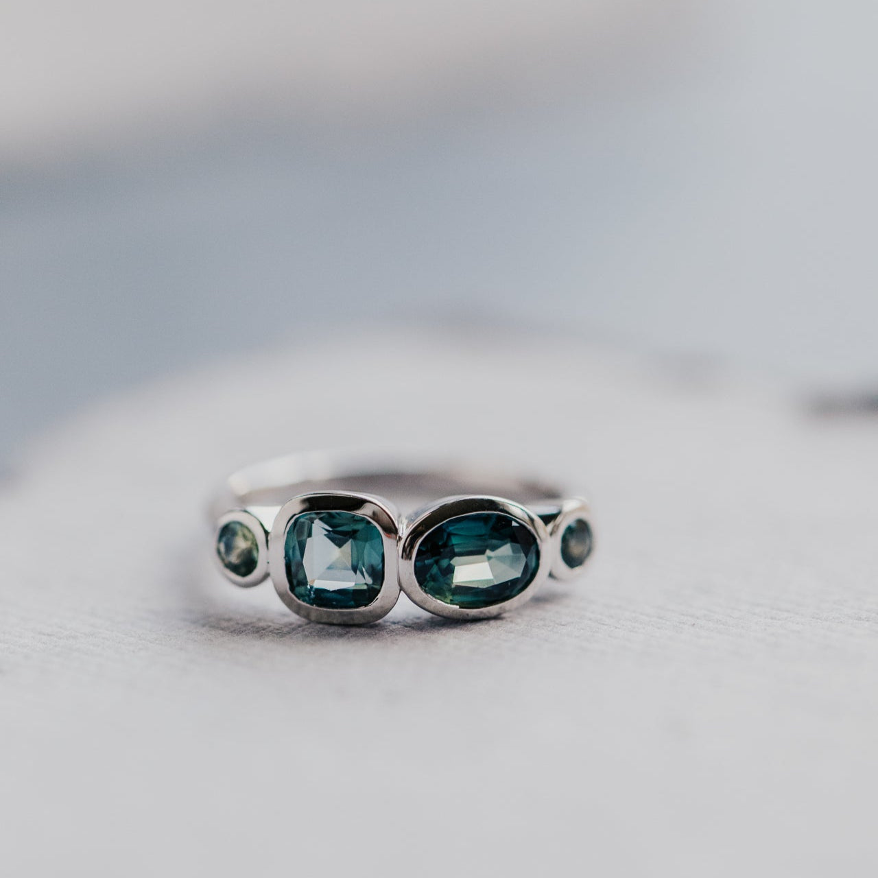 White gold ring with multiple green and blue sapphires  on a light gray background