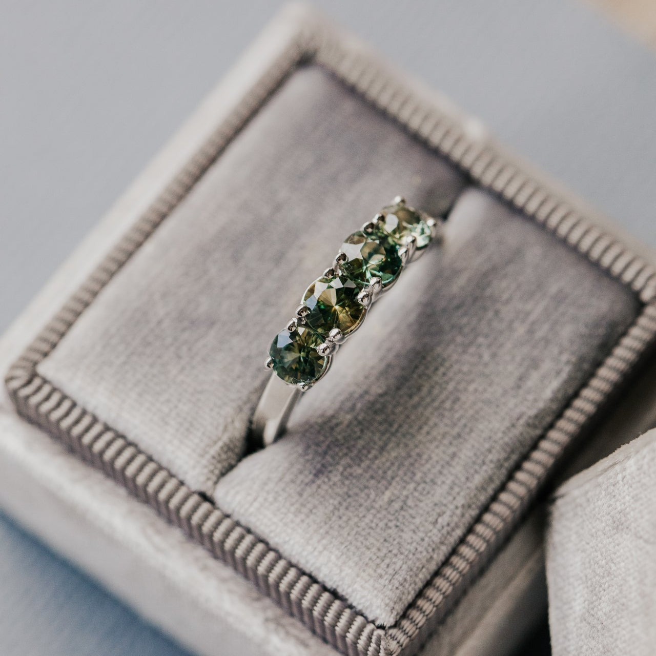 White gold ring with green and yellow sapphires on a gray cushioned jewelry box