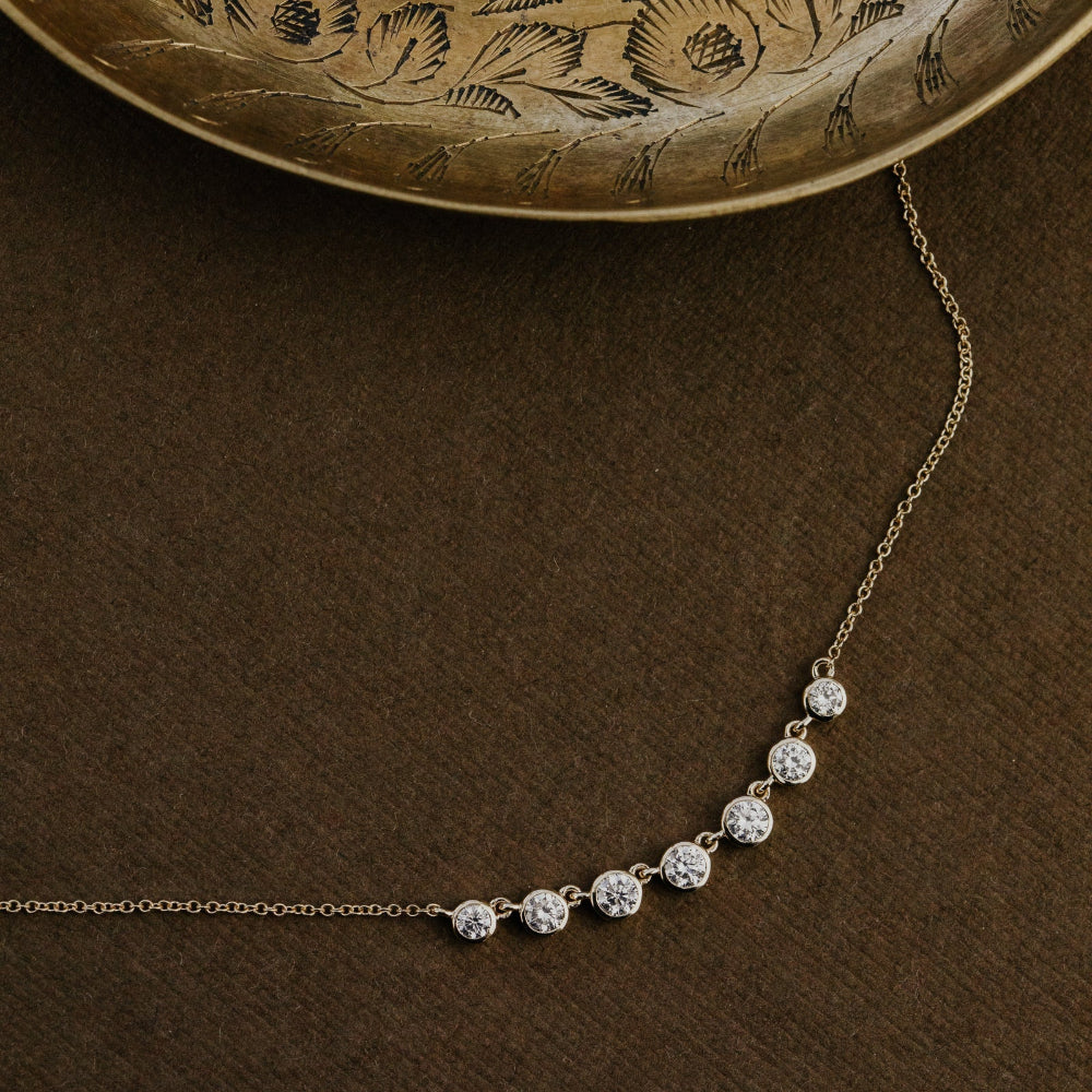 Gold necklace with seven round lab grown diamonds  on a brown surface with an embossed gold plate in the background.