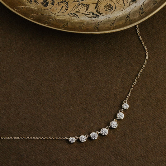 Gold necklace with seven round lab grown diamonds  on a brown surface with an embossed gold plate in the background.