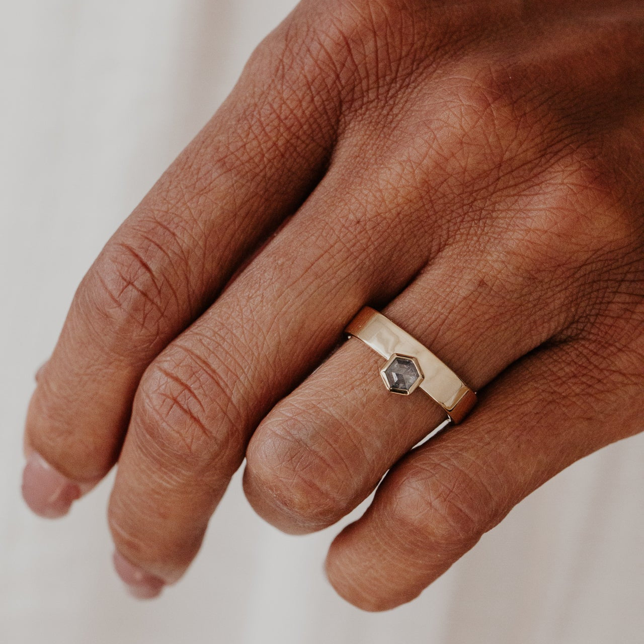 Hand wearing a gold ring with a gray hexagon rustic diamond on a neutral background.