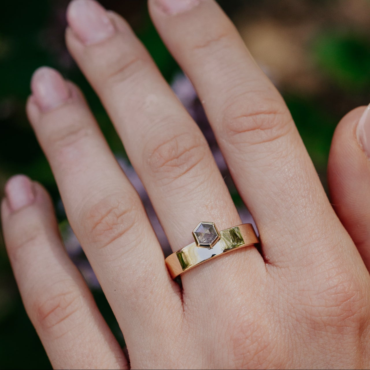 Hand wearing a gold ring with a hexagonal gray rustic diamond against a blurred natural background.