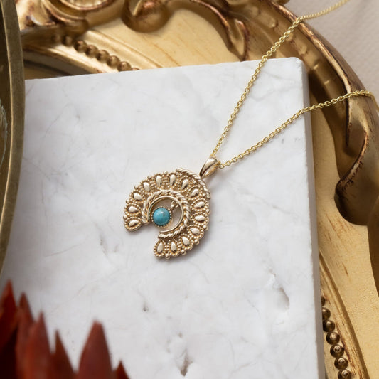 A gold necklace with a textured squash blossom pendant featuring a 3mm round turquoise stone, displayed on an adjustable chain, presented against a white marble background with a golden frame and a red flower.
